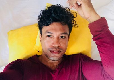 solo traveler wearing a purple sweater taking a selfie laying on the Butterfly Pea hotel room bed with yellow pillows